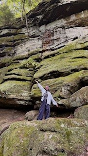 Nadia standing on a mossy rock formation outdoors with an arm raised
